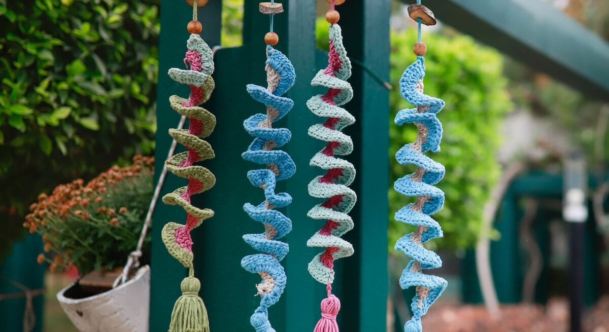 Bunte gehäkelte Spiralgirlanden mit Quasten hängen an einem Ast vor grünem Hintergrund mit Pflanzen und Holzstruktur.