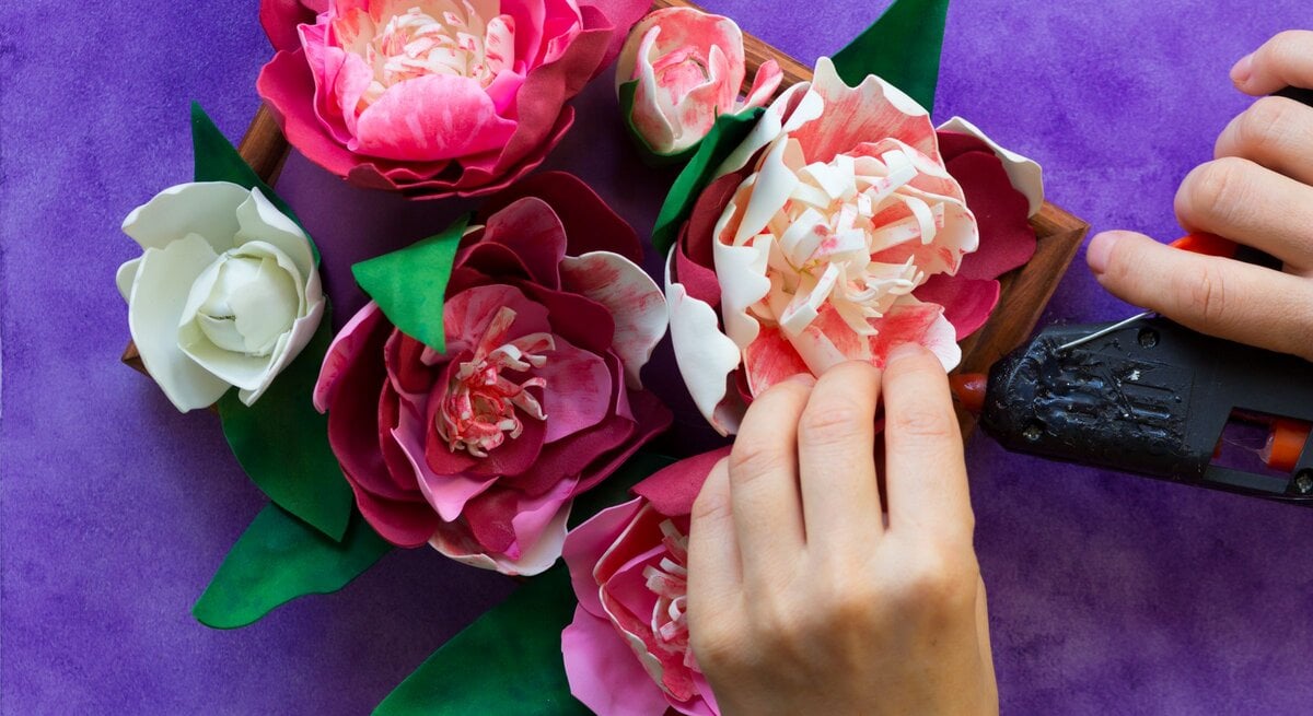 Hände gestalten rosa-weiße Pfingstrosen aus Watteads mit Heißklebepistole in einer Holzschale auf violettem Basteluntergrund. | © GettyImages/Tatiana Foxy