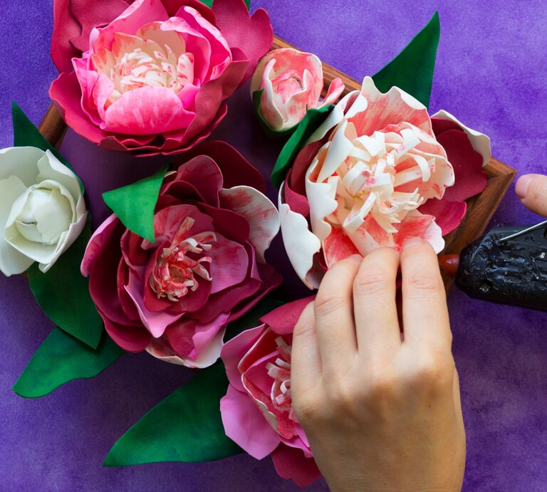 Hände gestalten rosa-weiße Pfingstrosen aus Watteads mit Heißklebepistole in einer Holzschale auf violettem Basteluntergrund. | © GettyImages/Tatiana Foxy