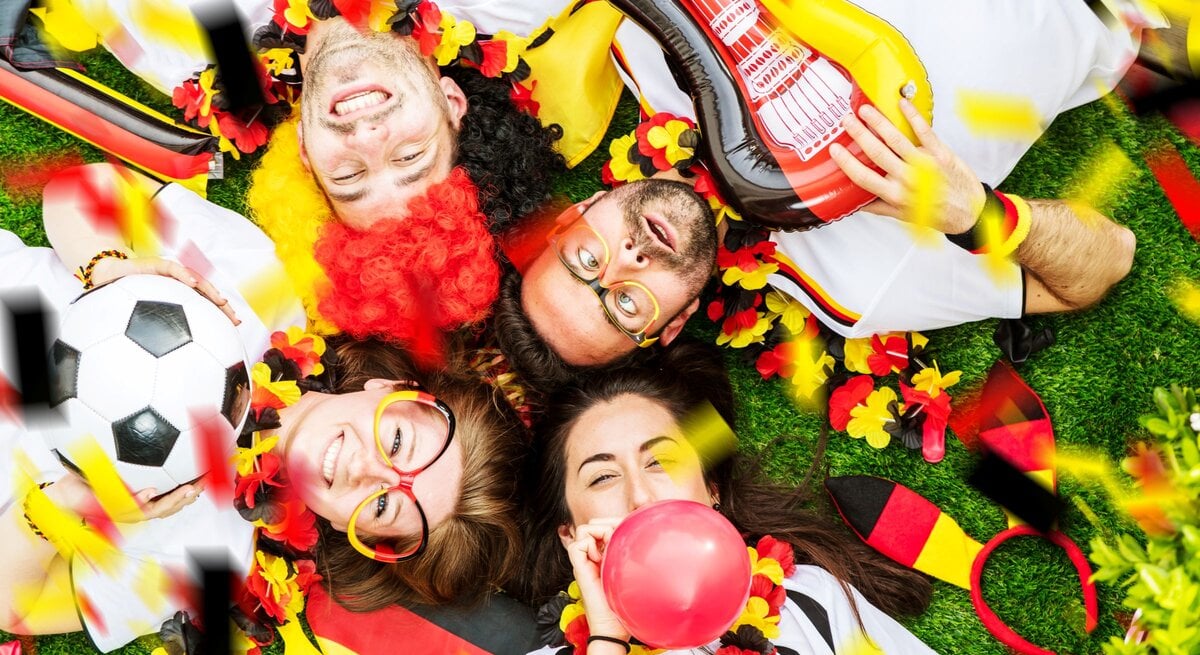 Vier jubelnde Fußballfans in Deutschlandfarben liegen lachend auf Rasen, umgeben von Konfetti, Ballons, Fahnen und Fußball.