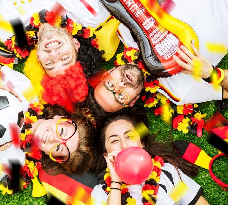 Vier jubelnde Fußballfans in Deutschlandfarben liegen lachend auf Rasen, umgeben von Konfetti, Ballons, Fahnen und Fußball. | © Shutterstock/KarepaStock