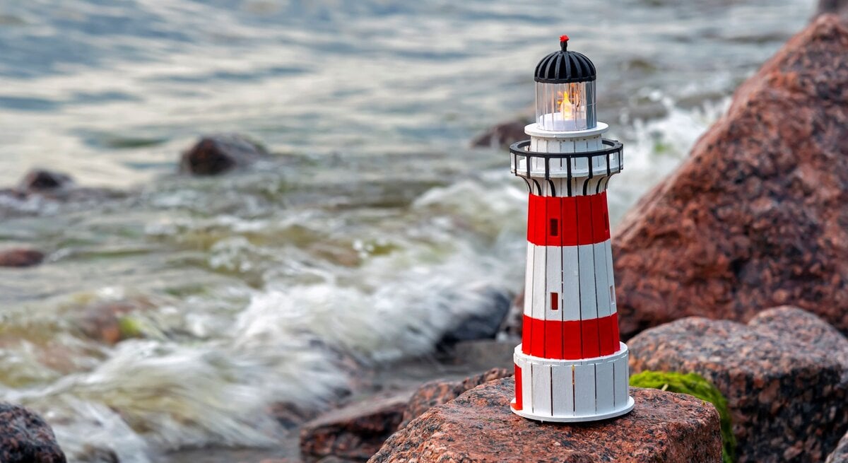Rot-weiß gestreifter Leuchtturm mit leuchtender Laterne steht auf Küstenfelsen, während sanfte Wellen im Hintergrund brechen.