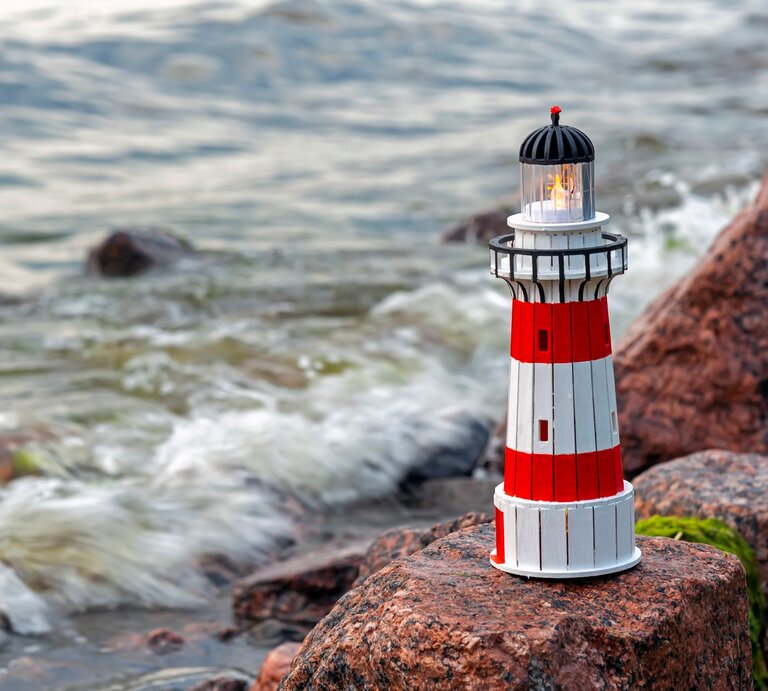 Rot-weiß gestreifter Leuchtturm mit leuchtender Laterne steht auf Küstenfelsen, während sanfte Wellen im Hintergrund brechen. | © GettyImages/Alexander Bondarenko / 500px