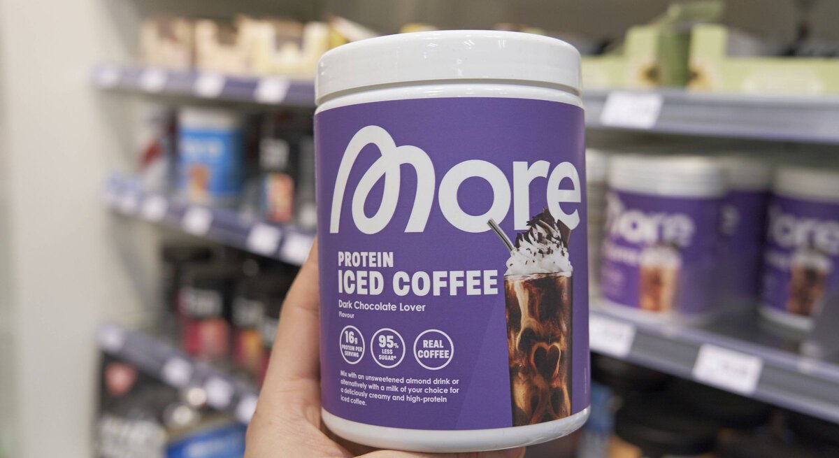 Eine Hand hält eine violette Dose Protein-Eiskaffee mit Schokoladengeschmack vor einem unscharfen Regal für Sportnahrung.