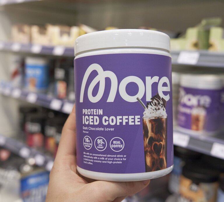 Eine Hand hält eine violette Dose Protein-Eiskaffee mit Schokoladengeschmack vor einem unscharfen Regal für Sportnahrung. | ©  IMAGO / imagebroker