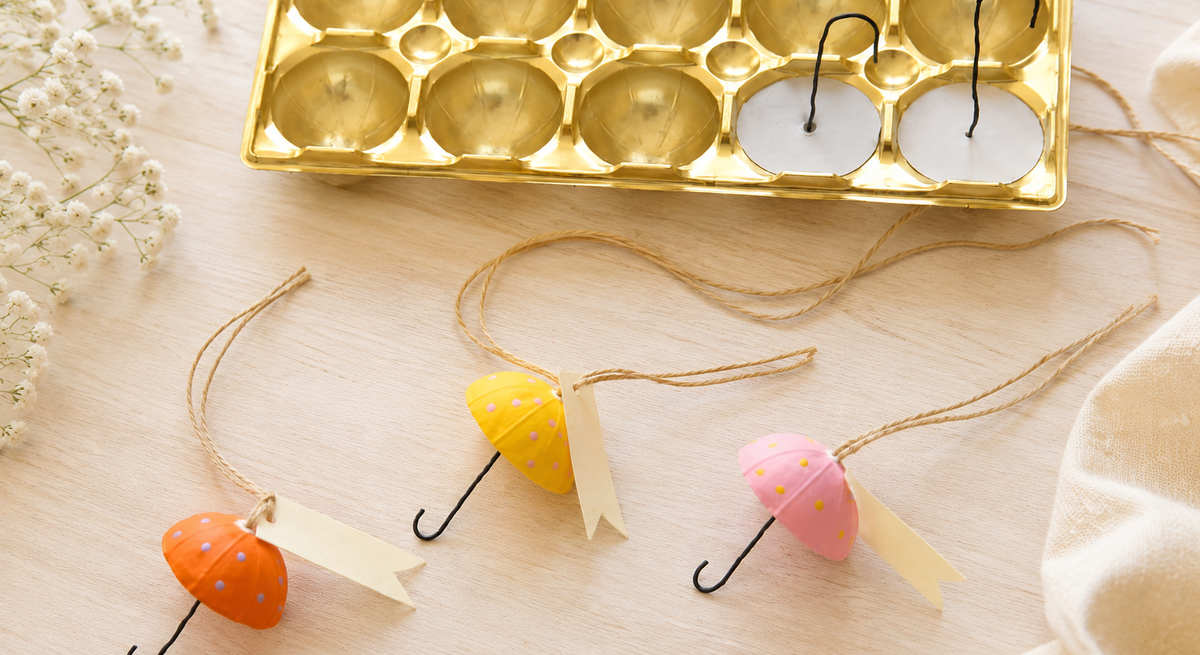 Selbst gebastelte Regenschirm-Anhänger in Pastellfarben liegen zwischen Schleifenband, weißen Blüten und leerer Toffifee-Verpackung aus Gold.