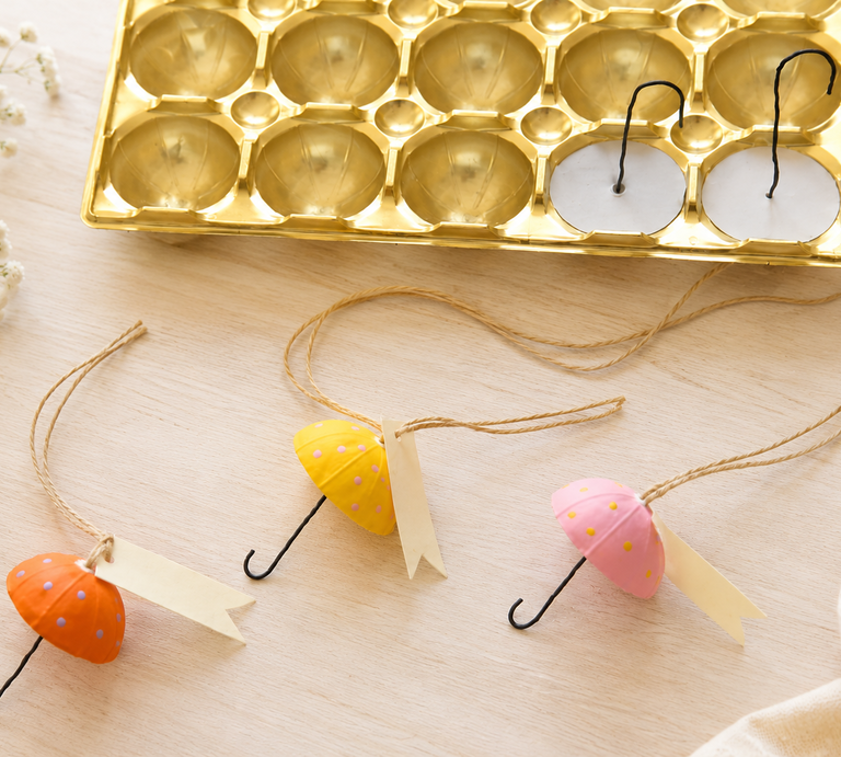 Selbst gebastelte Regenschirm-Anhänger in Pastellfarben liegen zwischen Schleifenband, weißen Blüten und leerer Toffifee-Verpackung aus Gold. | © ChatGPT