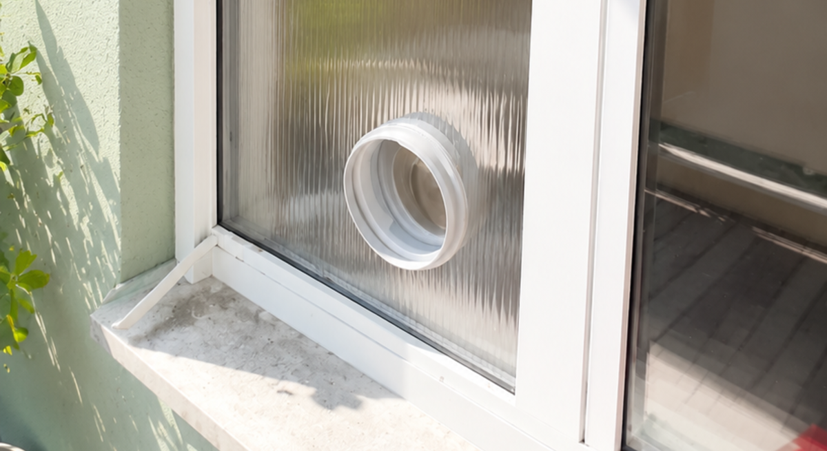 Milchige Stegplatte im weißen Fensterrahmen mit rundem Abluftstutzen; dahinter verschwimmen Bäume und Himmel im Sonnenlicht.