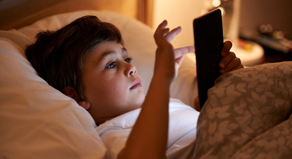 Ein braunhaariger Junge im Grundschulalter liegt abends im Bett und blickt konzentriert auf ein Amazon Kindle Colorsoft Kids in seinen Händen.