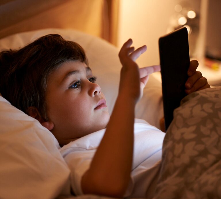 Ein braunhaariger Junge im Grundschulalter liegt abends im Bett und blickt konzentriert auf ein Amazon Kindle Colorsoft Kids in seinen Händen. | © Shutterstock/PeopleImages