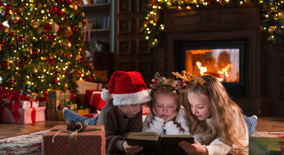 Drei Kinder lesen vertieft ein Buch vor dem Kamin, umgeben von Geschenken und festlich geschmücktem Weihnachtsbaum.