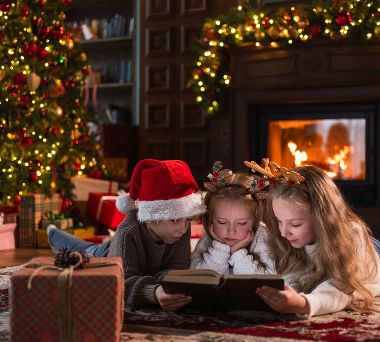 Drei Kinder lesen vertieft ein Buch vor dem Kamin, umgeben von Geschenken und festlich geschmücktem Weihnachtsbaum. | © Shutterstock/ Julia Zavalishina