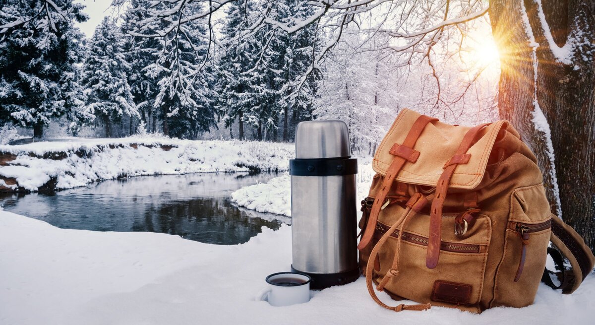 Rucksack und Thermoskanne stehen im Schnee vor winterlichem Wald und Fluss, Sonnenlicht fällt durch verschneite Äste.