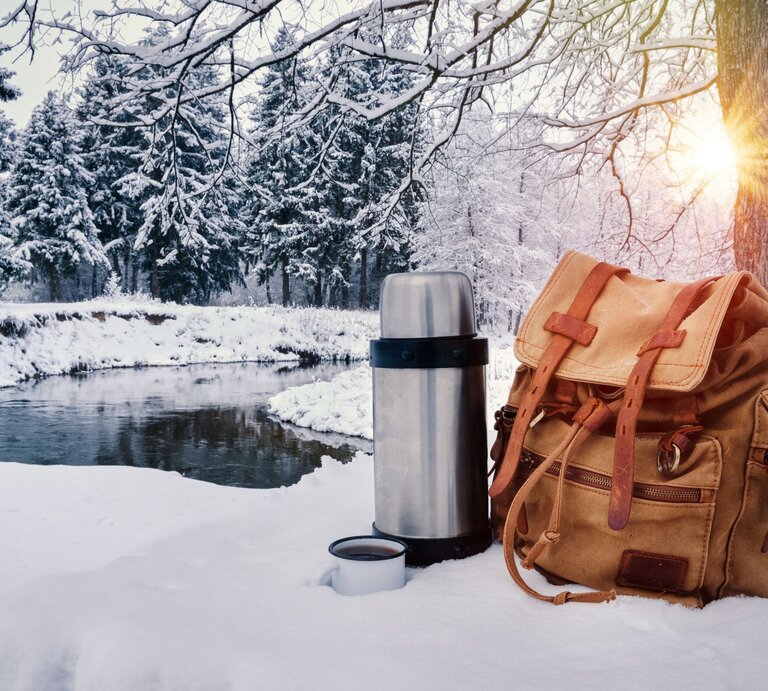 Rucksack und Thermoskanne stehen im Schnee vor winterlichem Wald und Fluss, Sonnenlicht fällt durch verschneite Äste. | © Shutterstock/ Pedal to the Stock