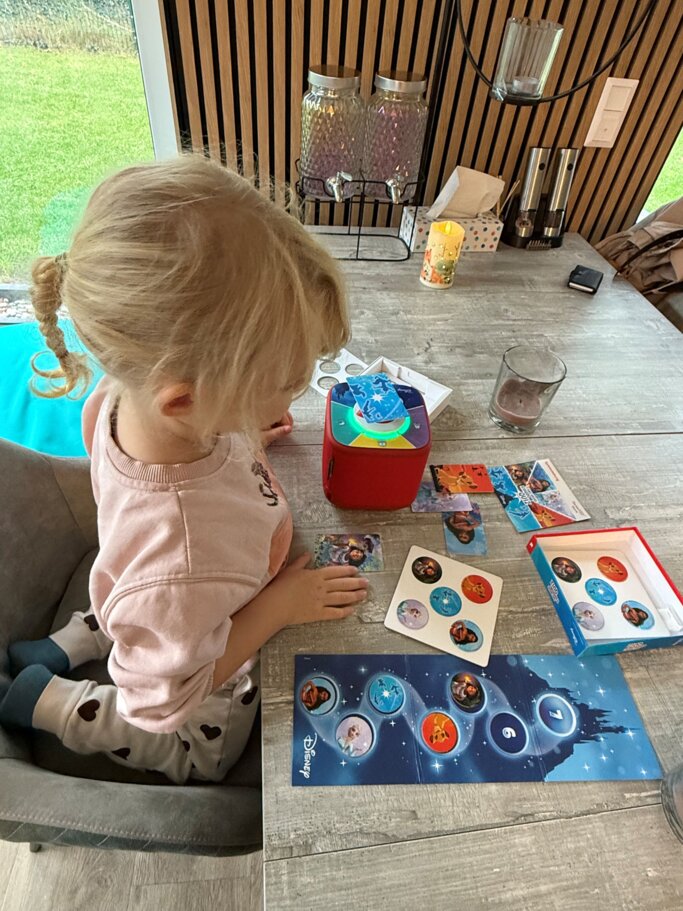Kleines blondes Mädchen im rosa Pulli spielt konzentriert mit Disney-Karten neben roter Toniebox an einem Holztisch. | © Madeleine Liedke 