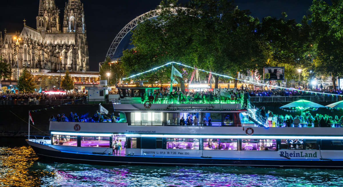 Zu sehen sind ein wunderschön beleuchtetes Boot mit dem Kölner Dom im Hintergrund