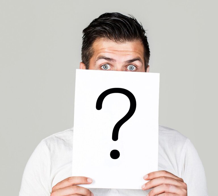 Ein junger Mann mit stechend blauen Augen hält ein Schild mit großem Fragezeichen vor sein Gesicht auf grauem Hintergrund. | © Adobe Stock / Yevhen