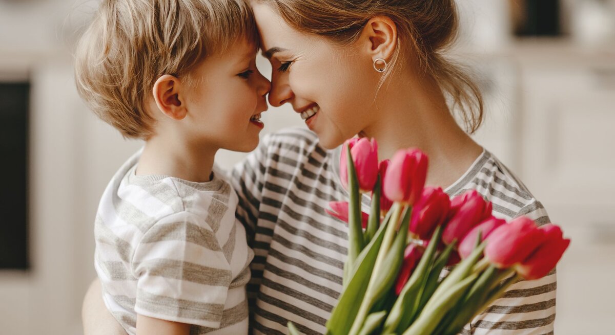 Eine Mutter nimmt ihren kleinen Sohn lächelnd in den Arm der ihr einen Strauß roter Tulpen schenkt. Man sieht beide von der Seite, wie sie sich anlächeln.