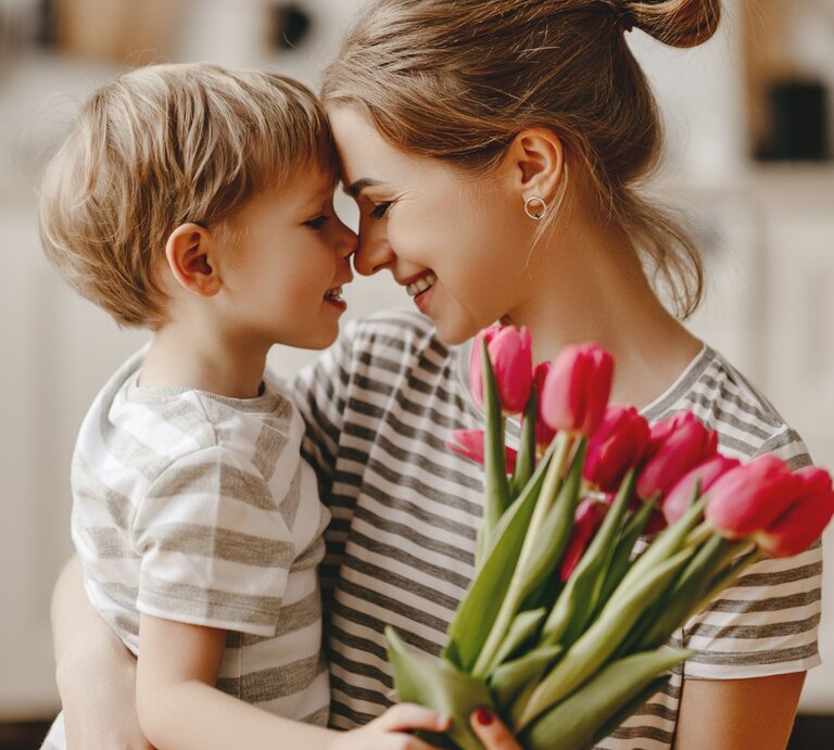 Eine Mutter nimmt ihren kleinen Sohn lächelnd in den Arm der ihr einen Strauß roter Tulpen schenkt. Man sieht beide von der Seite, wie sie sich anlächeln. | © gettyimages/ evgenyatamanenko