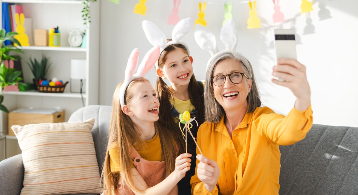 Ältere Frau mit grauen Haaren und Brille macht lachend ein Selfie mit zwei jungen Mädchen, alle tragen Hasenohren und Gelb.