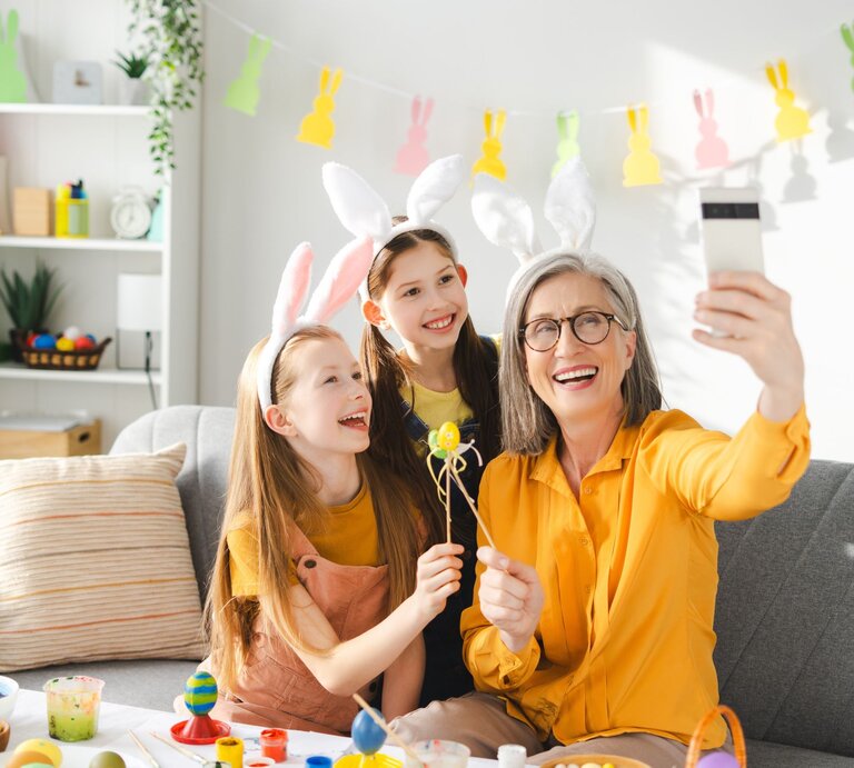 Ältere Frau mit grauen Haaren und Brille macht lachend ein Selfie mit zwei jungen Mädchen, alle tragen Hasenohren und Gelb. | © AdobeStock/Maria Vitkovska