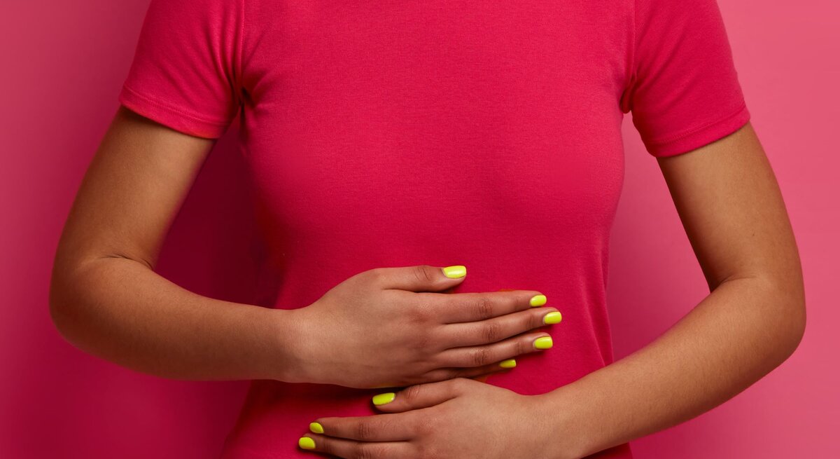Eine Frau in pinkem T-Shirt hält sich mit beiden Händen den Bauch, ihre gelb lackierten Fingernägel heben sich vom Hintergrund ab.