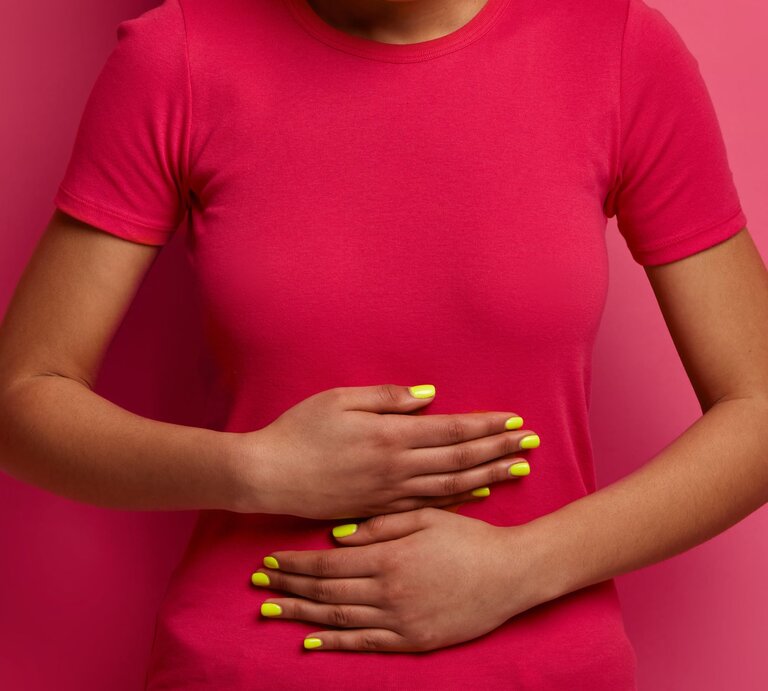 Eine Frau in pinkem T-Shirt hält sich mit beiden Händen den Bauch, ihre gelb lackierten Fingernägel heben sich vom Hintergrund ab. | © Adobe Stock / wayhome.studio
