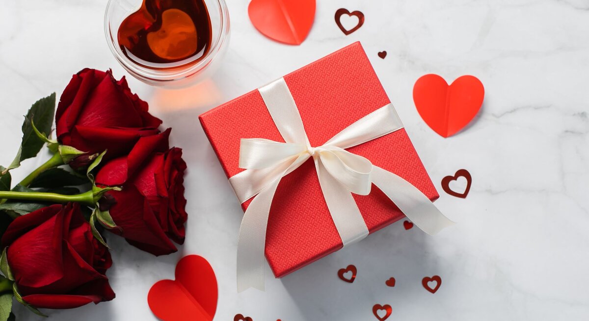 Drei rote Rosen liegen neben einer roten Geschenkbox mit Schleife, einer Tasse Tee in Herzform und Herzen auf hellem Marmortisch.