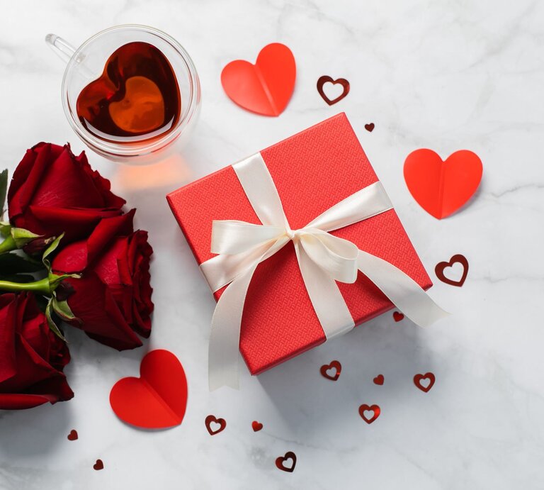 Drei rote Rosen liegen neben einer roten Geschenkbox mit Schleife, einer Tasse Tee in Herzform und Herzen auf hellem Marmortisch. | © GettyImages/Alexandr Kolesnikov