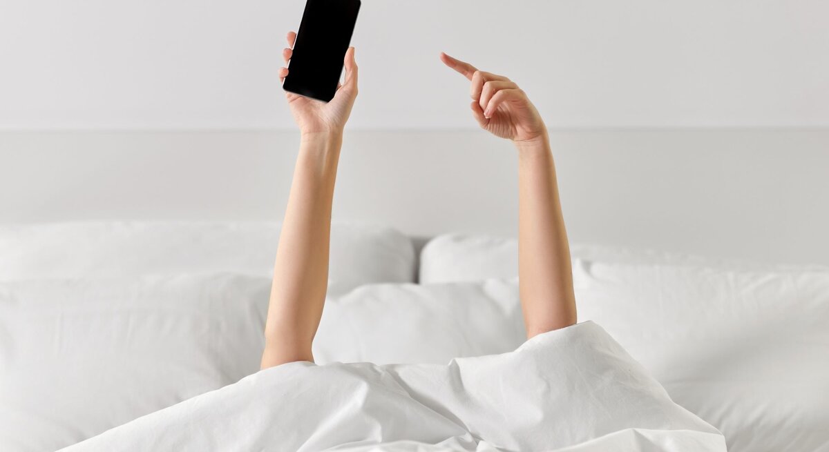 Zwei Arme strecken sich aus einer weißen Bettdecke, einer hält ein Smartphone, der andere zeigt mit dem Finger nach oben.