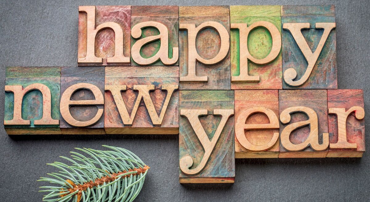 Bunte Holzbuchstaben formen den Schriftzug „Happy New Year“ auf einer grauen Unterlage, dekoriert mit einem grünen Tannenzweig.