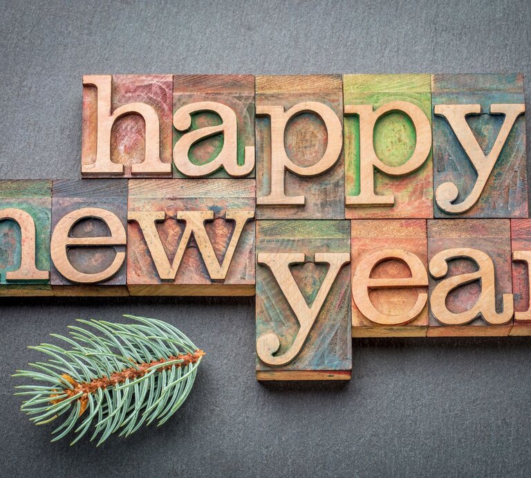 Bunte Holzbuchstaben formen den Schriftzug „Happy New Year“ auf einer grauen Unterlage, dekoriert mit einem grünen Tannenzweig. | © Adobe Stock/MarekPhotoDesign.com