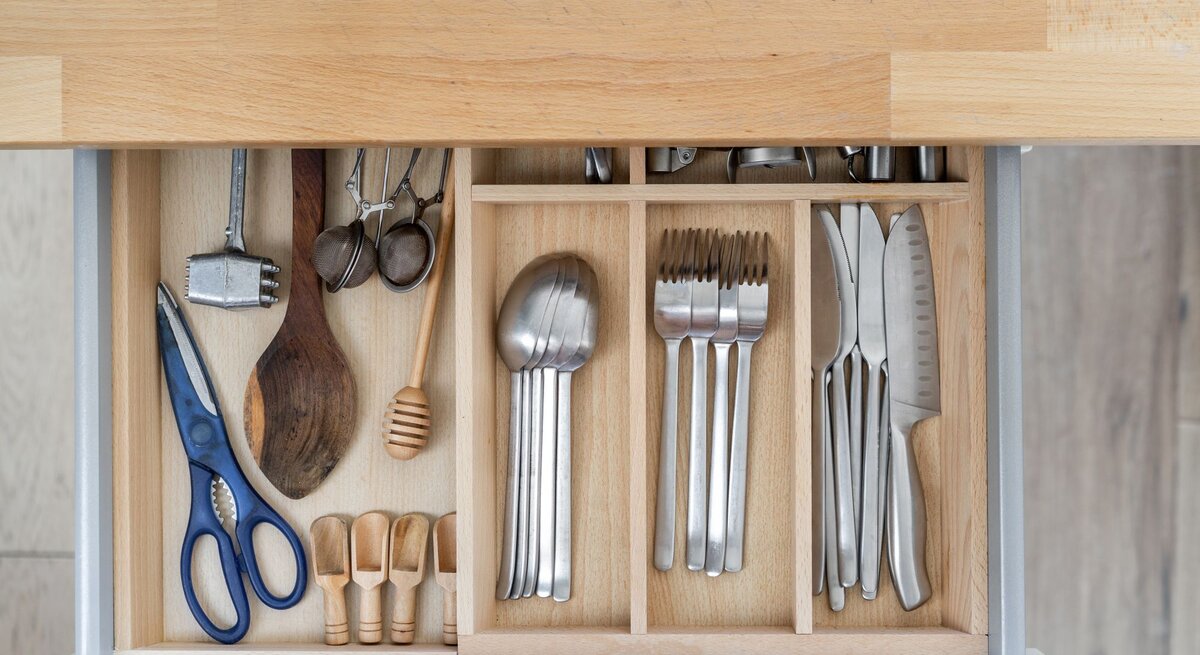 Geöffnete Besteckschublade mit einem Besteckkasten, gefüllt mit Besteck, Küchenutensilien wie Scheren, Holzlöffel und Messern auf einer hellen Arbeitsplatte.