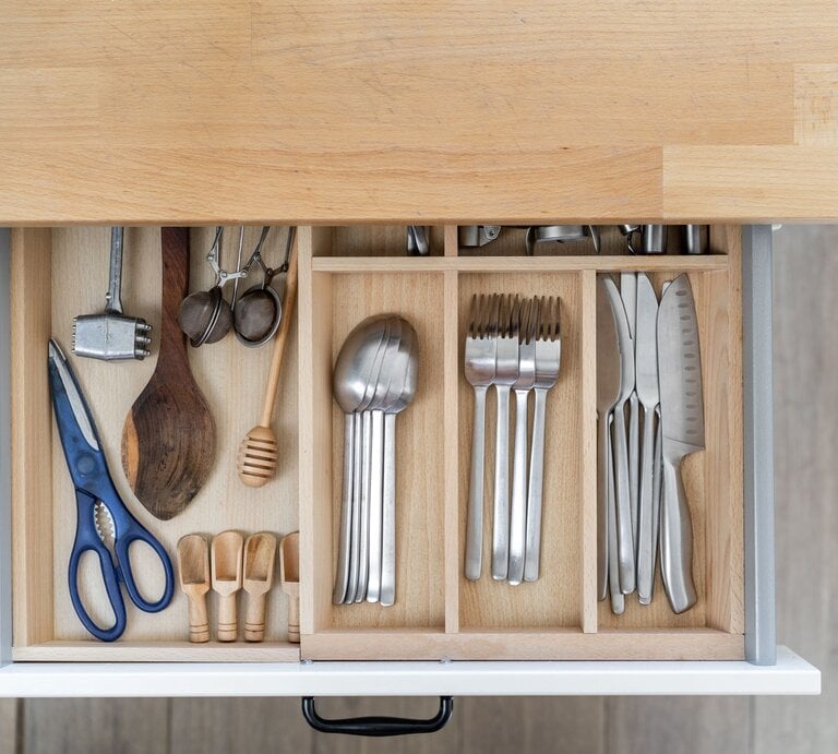 Geöffnete Besteckschublade mit einem Besteckkasten, gefüllt mit Besteck, Küchenutensilien wie Scheren, Holzlöffel und Messern auf einer hellen Arbeitsplatte. | © AdobeStock/ galaganov