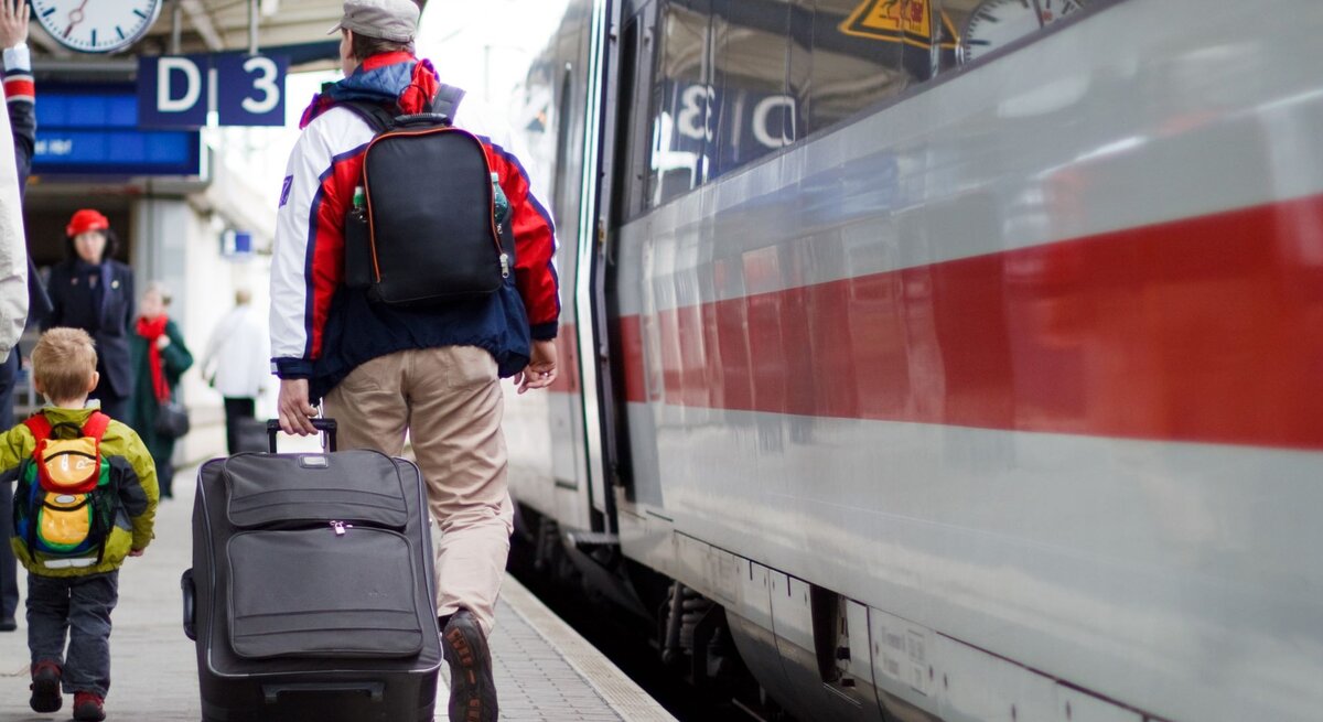Ein Mann mit grauer Mütze und roter Jacke zieht einen Koffer, begleitet von einem Jungen mit gelbem Rucksack, auf einem Bahnsteig, an dem ein ICE der Deutschen Bahn hält.