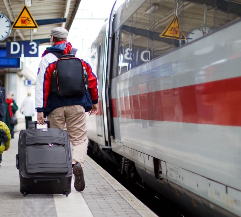 Ein Mann mit grauer Mütze und roter Jacke zieht einen Koffer, begleitet von einem Jungen mit gelbem Rucksack, auf einem Bahnsteig, an dem ein ICE der Deutschen Bahn hält. | © Adobe Stock/Henlisatho