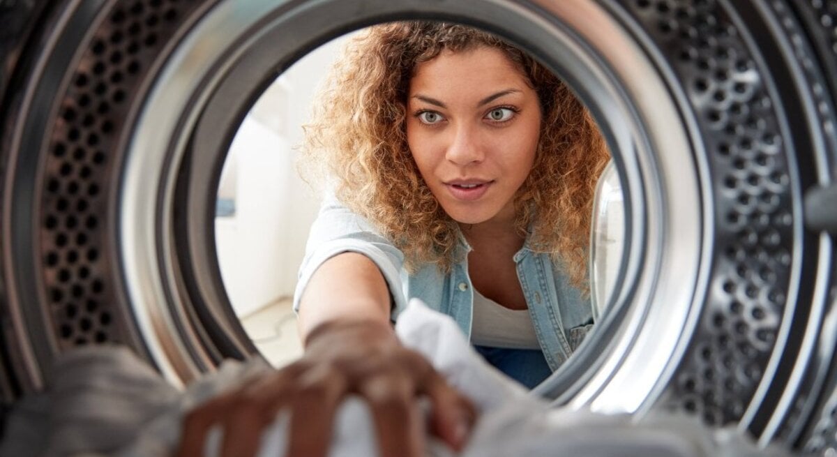 Junge Frau mit lockigem Haar und Denim-Hemd greift konzentriert in die Trommel einer Waschmaschine, aus deren Innerem fotografiert.