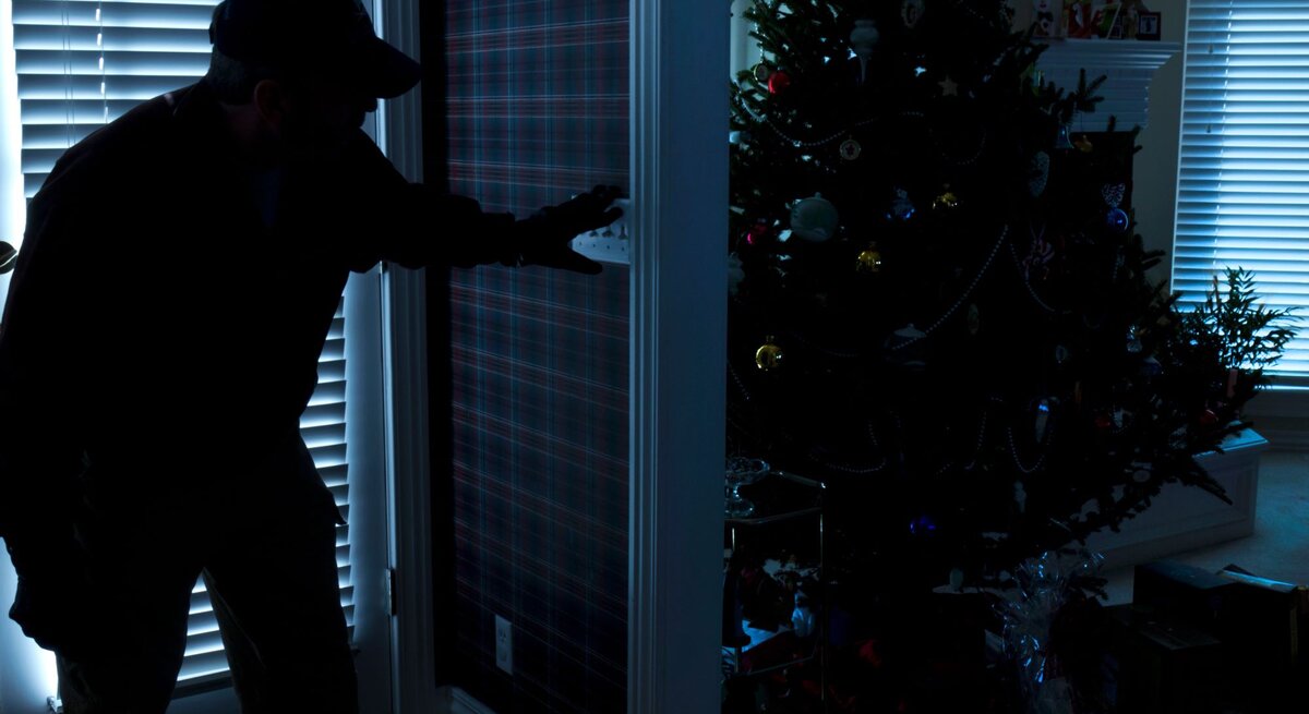 Überwachungskameras können hiervor schützen: Silhouette eines Einbrechers, der vorsichtig eine Tür öffnet, während im Hintergrund ein festlich dekorierter Weihnachtsbaum steht.