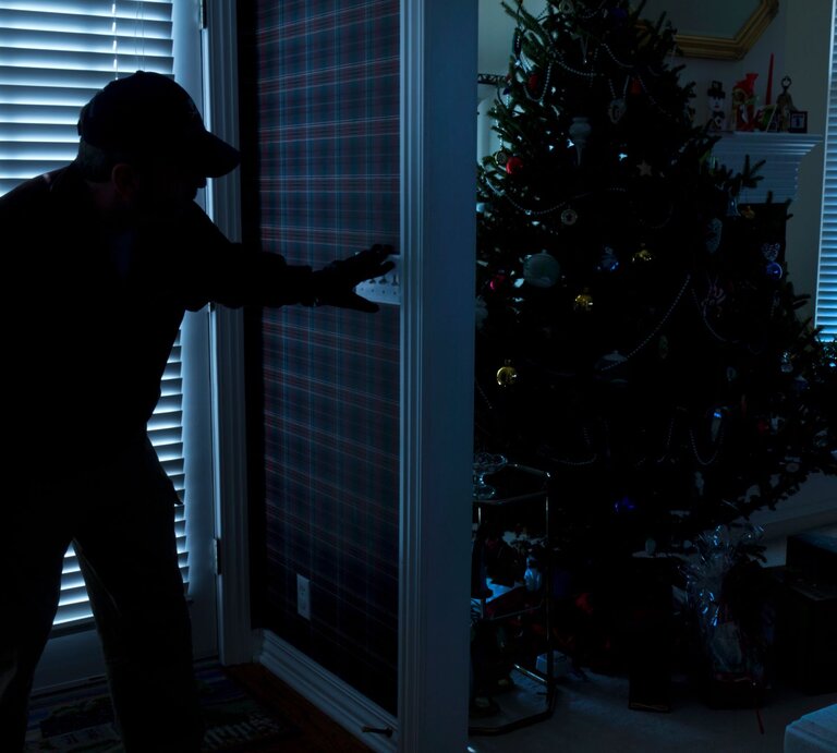 Überwachungskameras können hiervor schützen: Silhouette eines Einbrechers, der vorsichtig eine Tür öffnet, während im Hintergrund ein festlich dekorierter Weihnachtsbaum steht. | © AdobeStock/ricardoreitmeyer