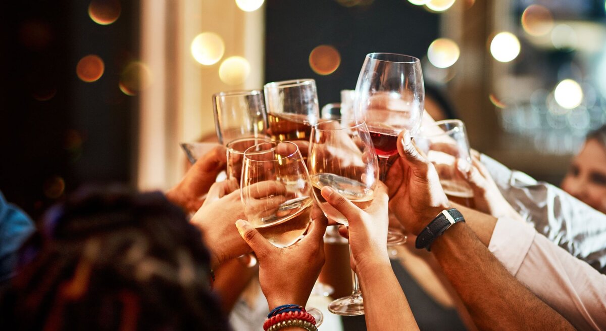 Mehrere Hände heben Gläser mit Wein und Sekt für einen fröhlichen Toast vor einem Hintergrund aus warmen Lichtern.