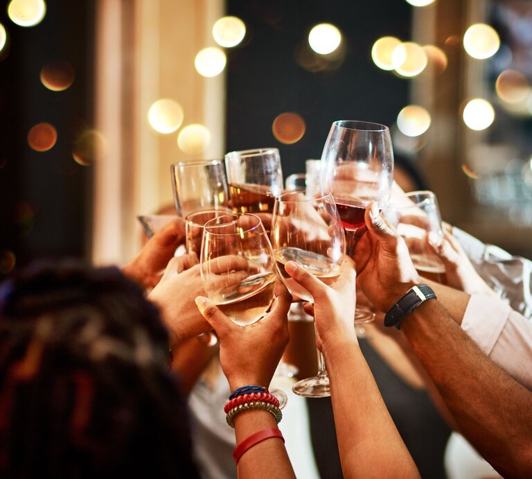 Mehrere Hände heben Gläser mit Wein und Sekt für einen fröhlichen Toast vor einem Hintergrund aus warmen Lichtern. | © AdobeStock/Malambo/peopleimages.com