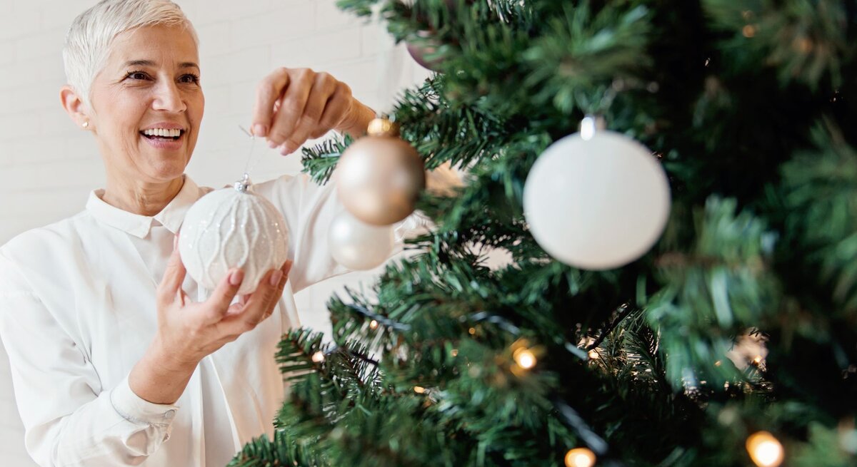 Eine lächelnde ältere Frau mit kurzen grauen Haaren hängt eine weiße Christbaumkugel an einen festlich geschmückten Tannenbaum.