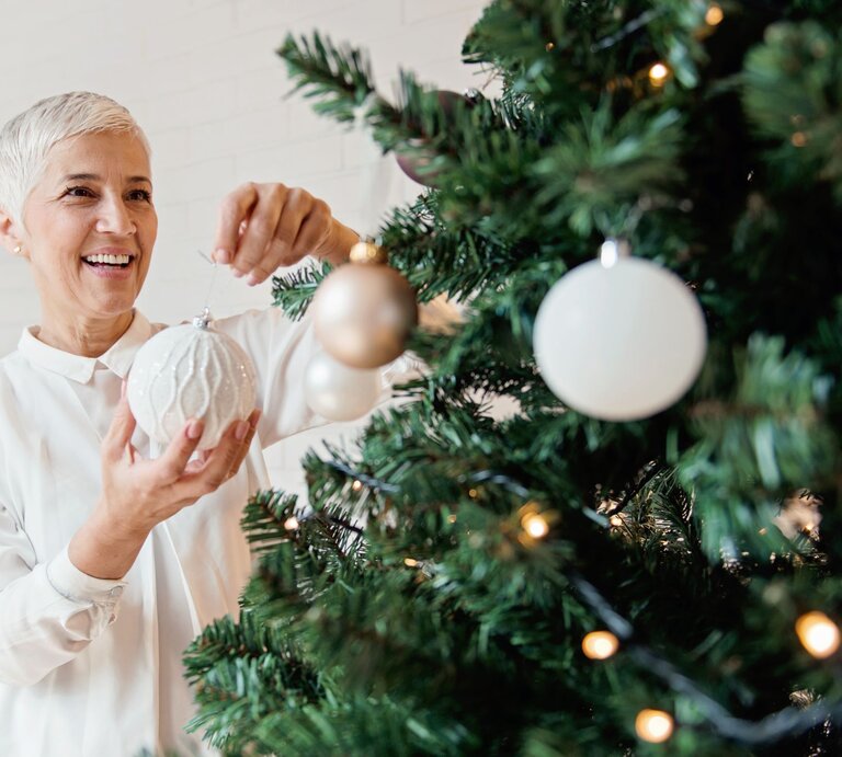 Eine lächelnde ältere Frau mit kurzen grauen Haaren hängt eine weiße Christbaumkugel an einen festlich geschmückten Tannenbaum. | © Adobe Stock/Tijana
