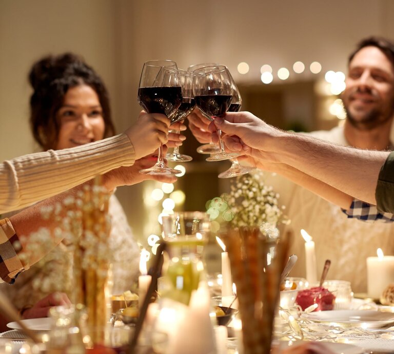 Vier Personen stoßen mit Rotweingläsern zu Weihnachten an, lächelnd an einem festlich gedeckten Tisch mit Kerzenlicht und Dekoration. | © Adobe Stock/Syda Productions