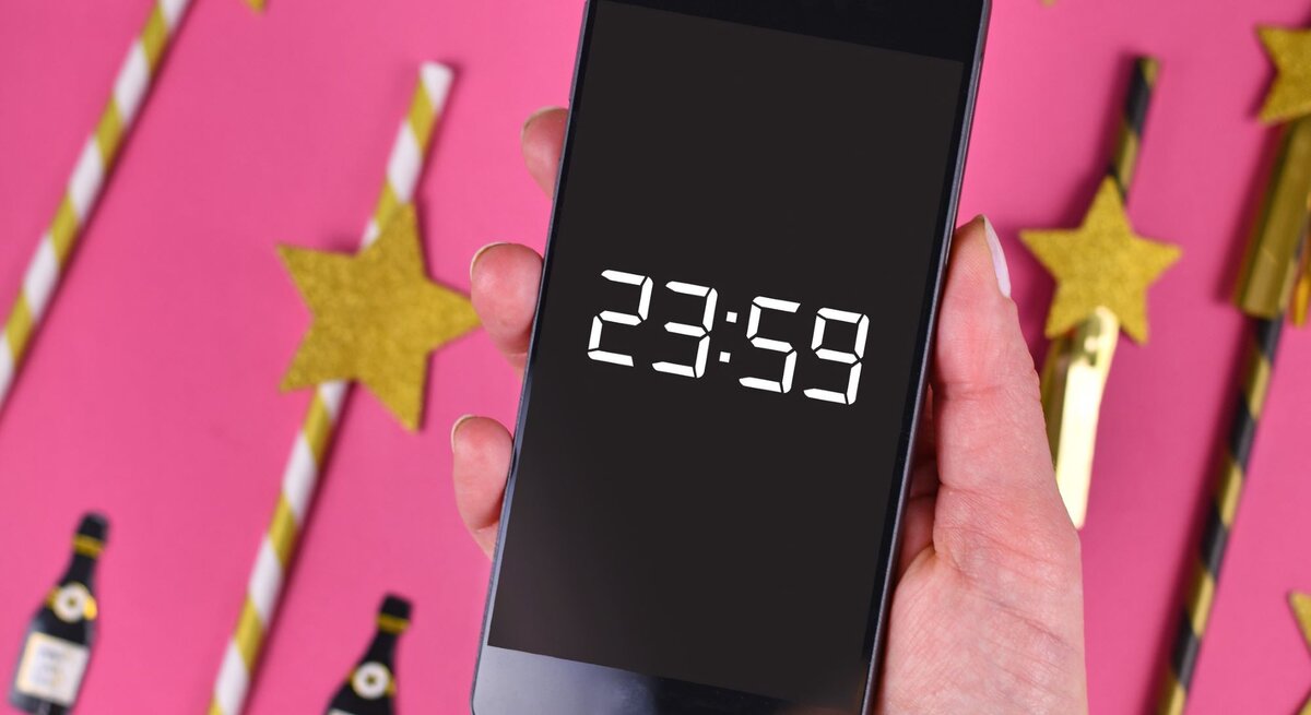Silvestersprüche: Eine Hand mit rosa lackierten Nägeln hält ein Smartphone mit 23:59-Anzeige, umgeben von goldenen Sternen und Partydekorationen auf pinkem Hintergrund.