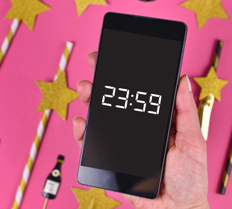 Silvestersprüche: Eine Hand mit rosa lackierten Nägeln hält ein Smartphone mit 23:59-Anzeige, umgeben von goldenen Sternen und Partydekorationen auf pinkem Hintergrund. | © AdobeStock/Firn