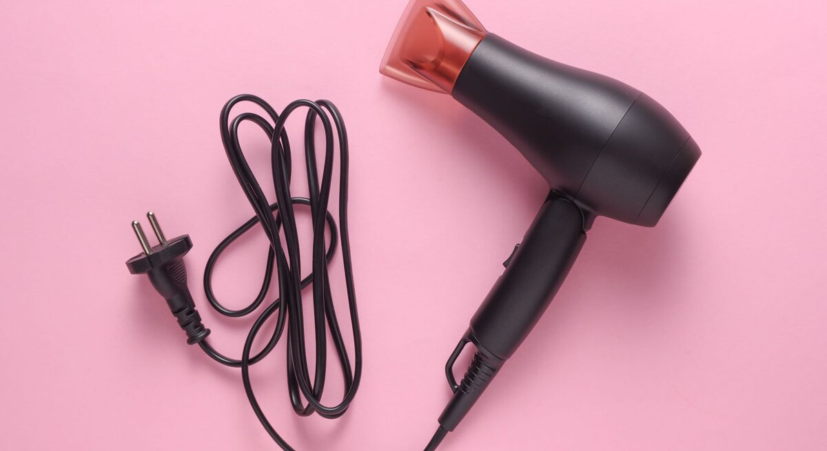 Schwarzer Haartrockner mit langem Kabel und europäischem Stecker liegt auf hellem rosa Hintergrund.
