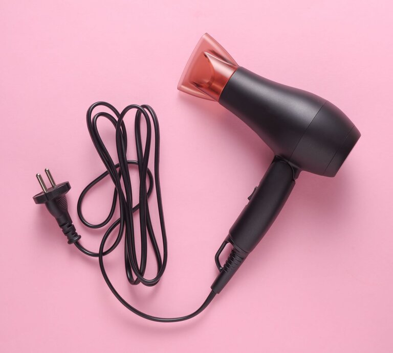 Schwarzer Haartrockner mit langem Kabel und europäischem Stecker liegt auf hellem rosa Hintergrund. | © Adobe Stock/splitov27