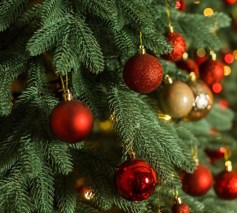 Nahaufnahme eines geschmückten künstlichen Weihnachtsbaumes mit roten und goldenen Weihnachtskugeln vor unscharfem, warm beleuchtetem Hintergrund. | © AdobeStock/ Oleksandr Tsybulskyy