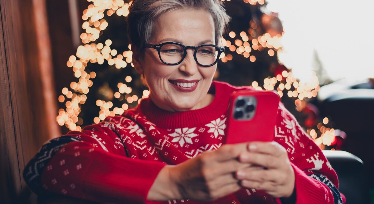 Eine grauhaarige Frau mittleren Alters mit Brille lächelt, während sie auf einem Sessel in rotem Weihnachtspullover ein Smartphone hält.